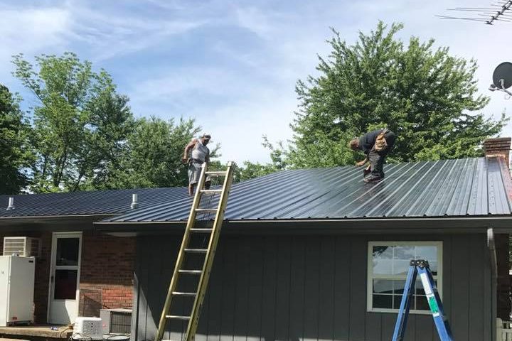 Two roofers installing dark metal roofing on a gray-sided house, with extension and step ladders — Kentucky