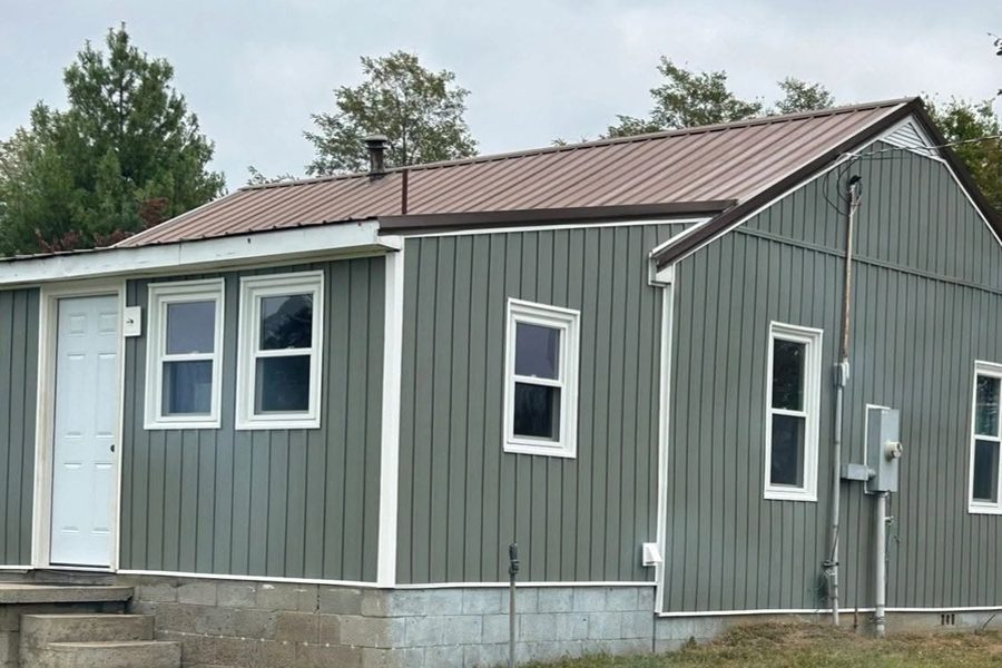 Small house finished with green vertical board-and-batten siding, white trim and a brown metal roof