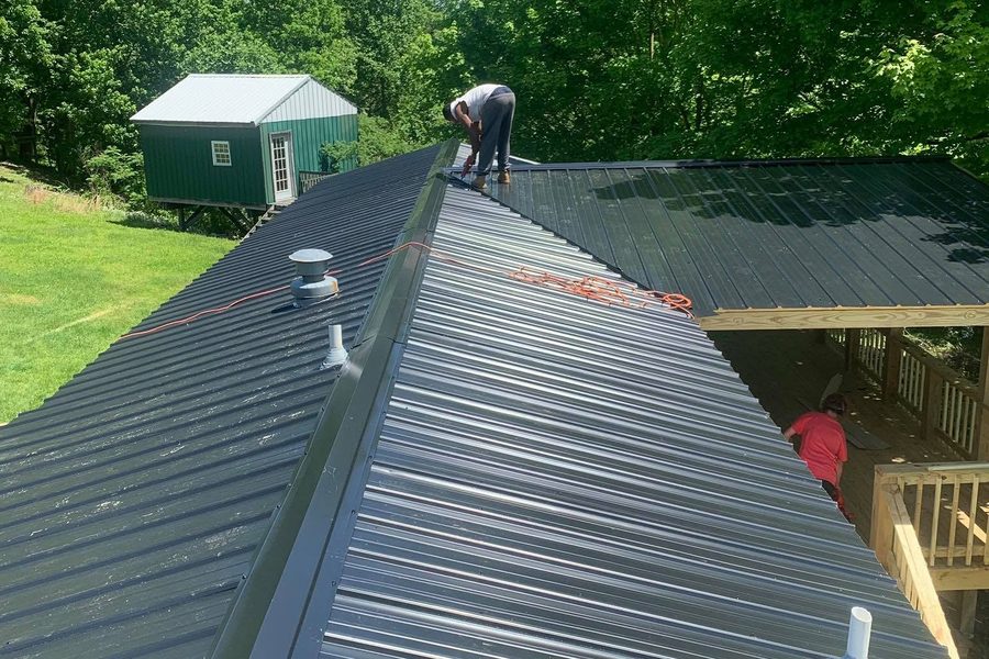 Two crew members installing dark green metal roofing on a house, with a new wood deck below