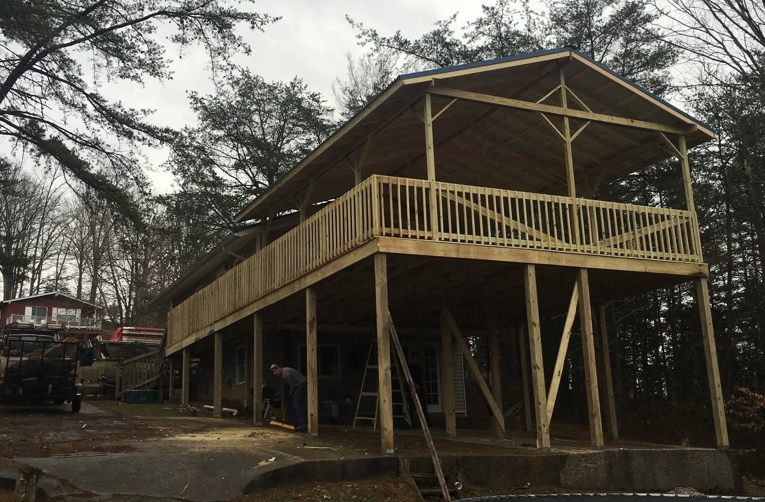Covered two-level wood deck on tall posts behind a wooded home, with a crew member working underneath