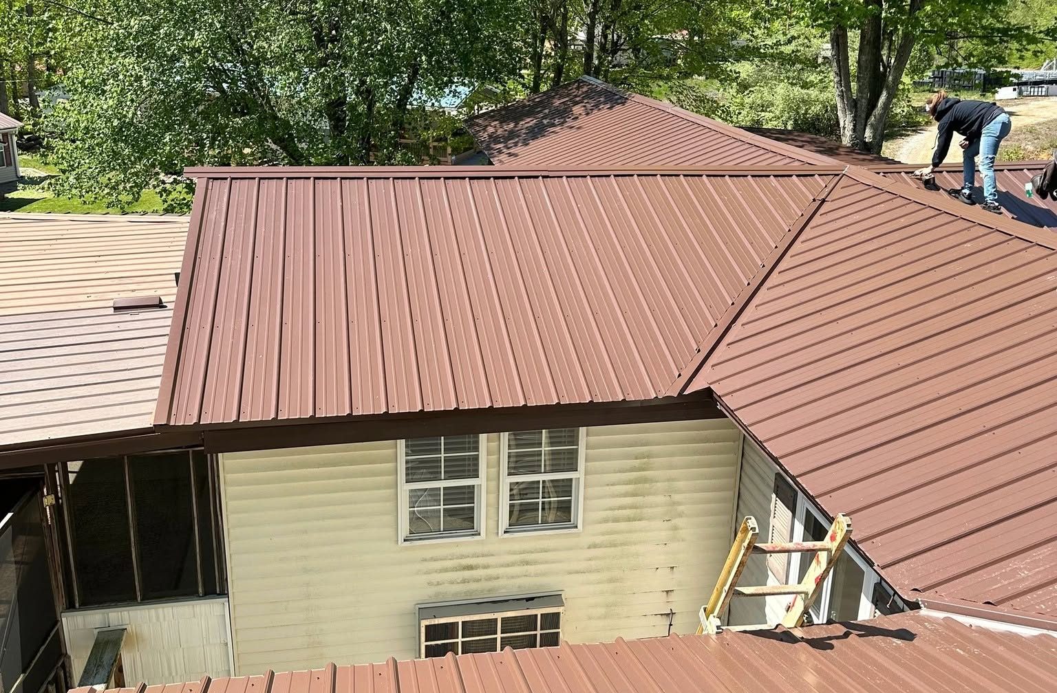 Crew member installing brown metal roofing on a two-story house with pale yellow siding