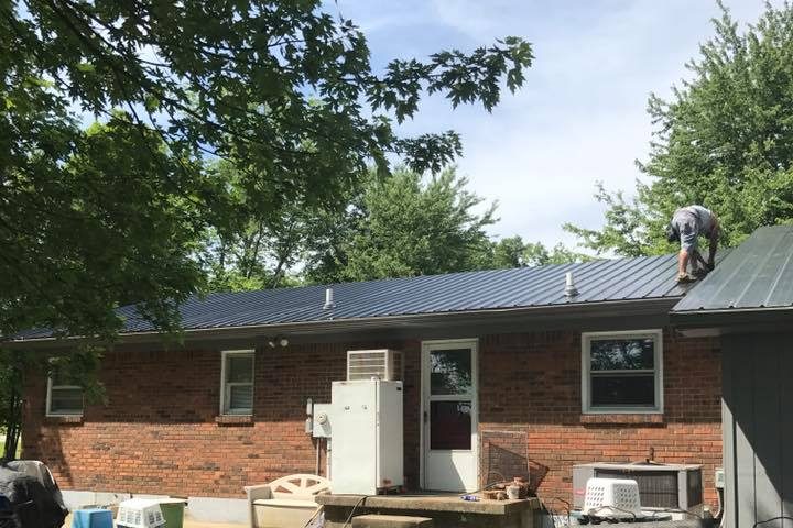 New dark metal roof going on a brick ranch house, with a roofer at the ridge — Kentucky